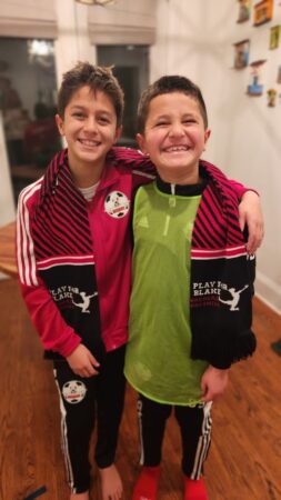 A photo of two boys modeling one knit 'Play for Blake' scarf over both their shoulders.
